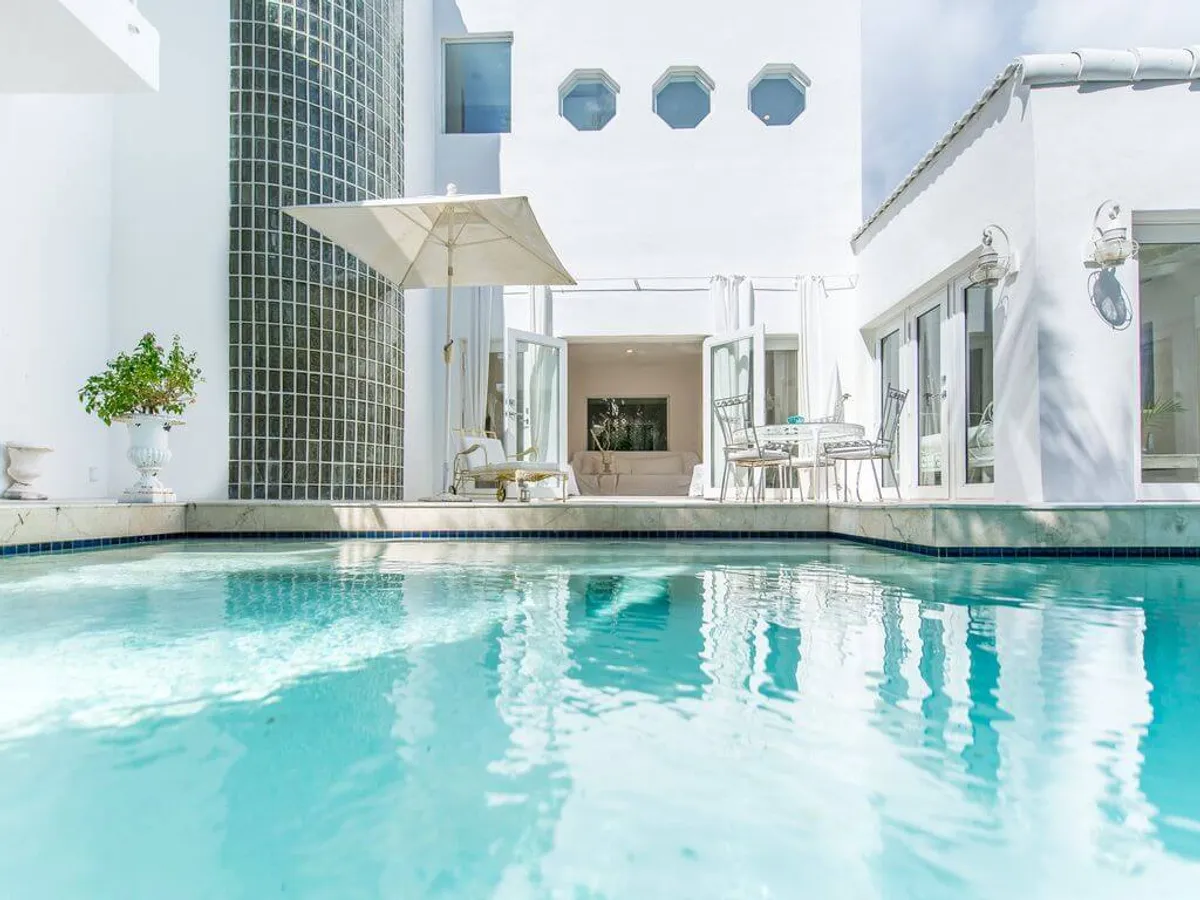 The hero image showcases a sparkling heated swimming pool surrounded by elegant outdoor patio furniture, with white French doors seamlessly connecting the sun-drenched poolside area to the stylish interior. Lush greenery and a unique glass block curved wall add a touch of modern sophistication, creating a tranquil, private oasis in the heart of Miami.