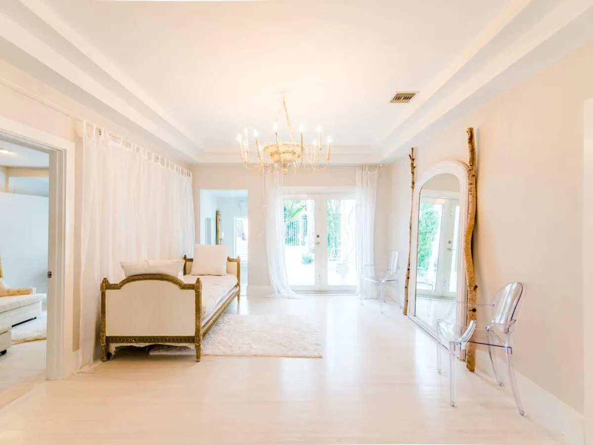 This image showcases an elegant, sunlit interior space featuring an ornate vintage settee with gold detailing, a large rustic wood-framed mirror, and a delicate chandelier. Sheer white curtains diffuse natural light, creating a tranquil atmosphere and offering a visual connection to the lush tropical exterior beyond the French doors.
