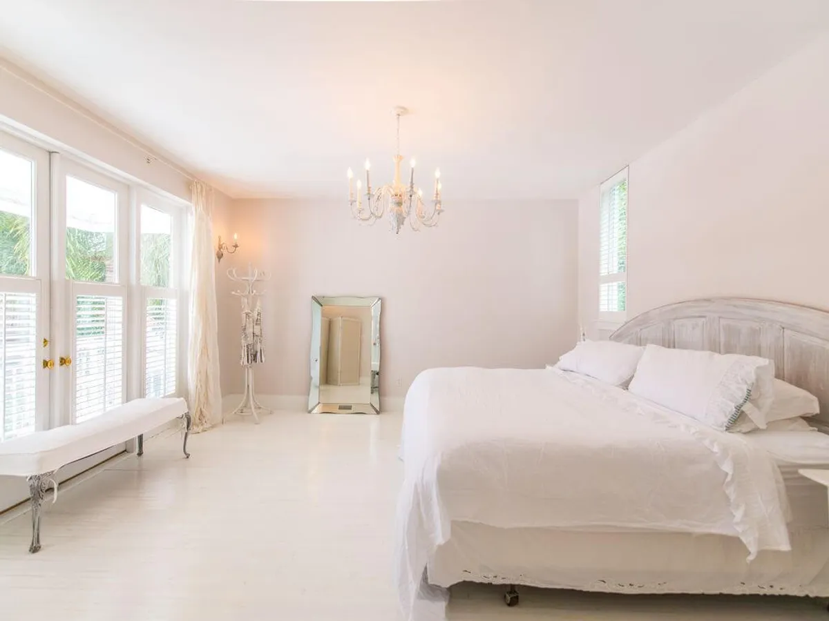 A spacious, sun-drenched bedroom with a large white bed adorned with textured bedding, a chandelier lighting fixture, a full-length mirror, and French doors with white blinds and sheer curtains. Natural light pours into the room, emphasizing its restful and inviting atmosphere.