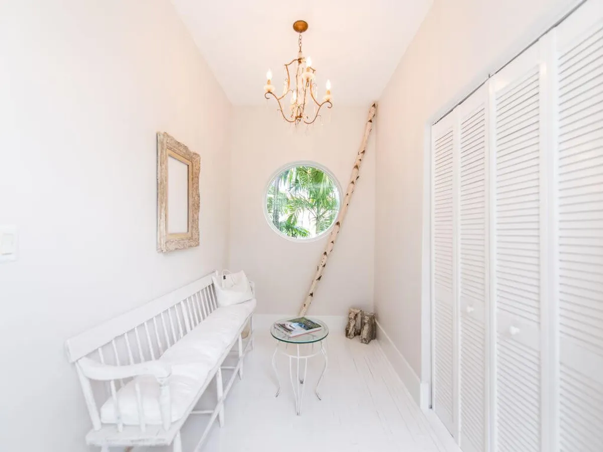 A cozy breakfast nook featuring a white cushioned bench, round glass-top table, and a unique circular window framing tropical greenery. Rustic details, such as a wooden ladder, add character, while the abundant natural light enhances the airy, welcoming feel ideal for morning coffee or intimate gatherings.