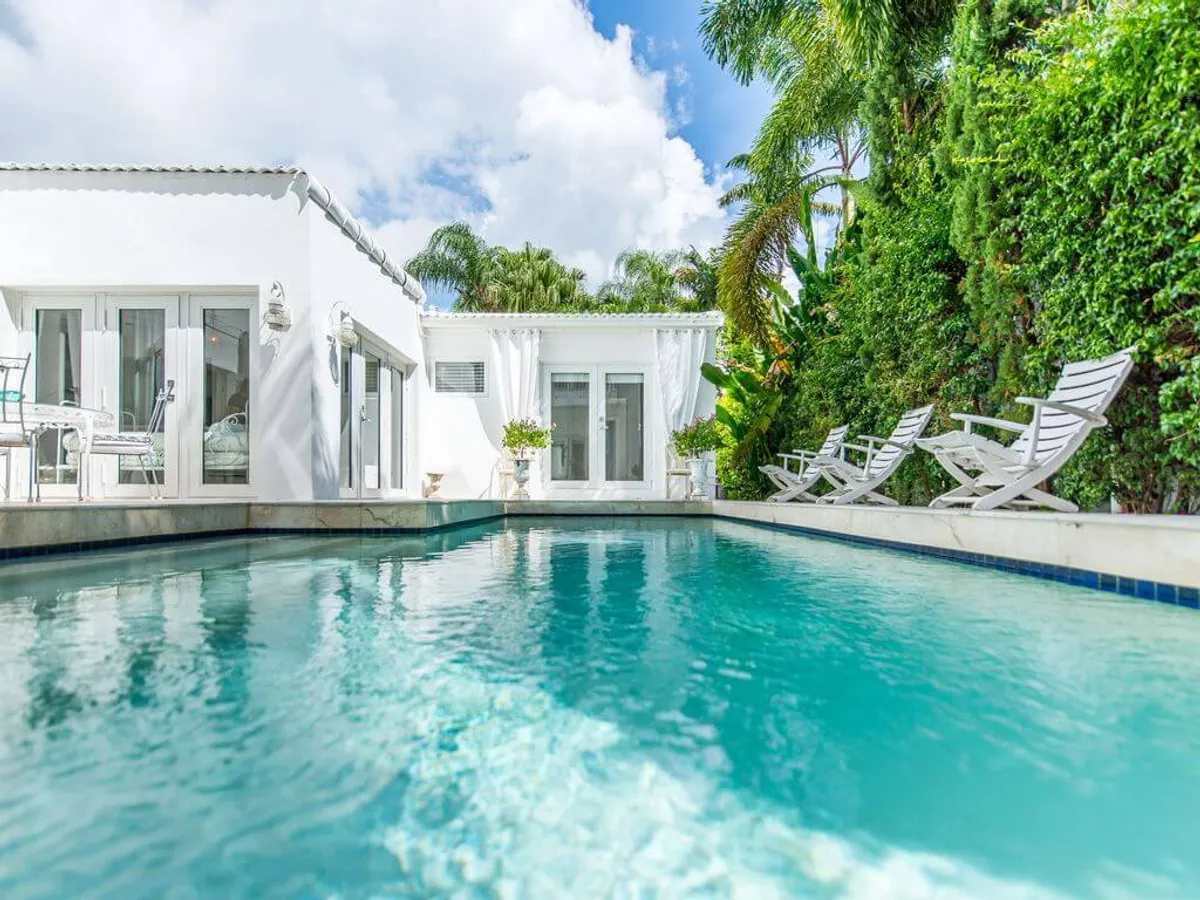 A sparkling blue swimming pool takes center stage, bordered by white lounge chairs perfectly aligned for sunbathing. The pool is surrounded by lush, green tropical plants and tall palms, creating a secluded oasis feel. The modern white house in the background features French doors and large windows that open directly onto the pool area, suggesting a seamless flow between indoor and outdoor living.