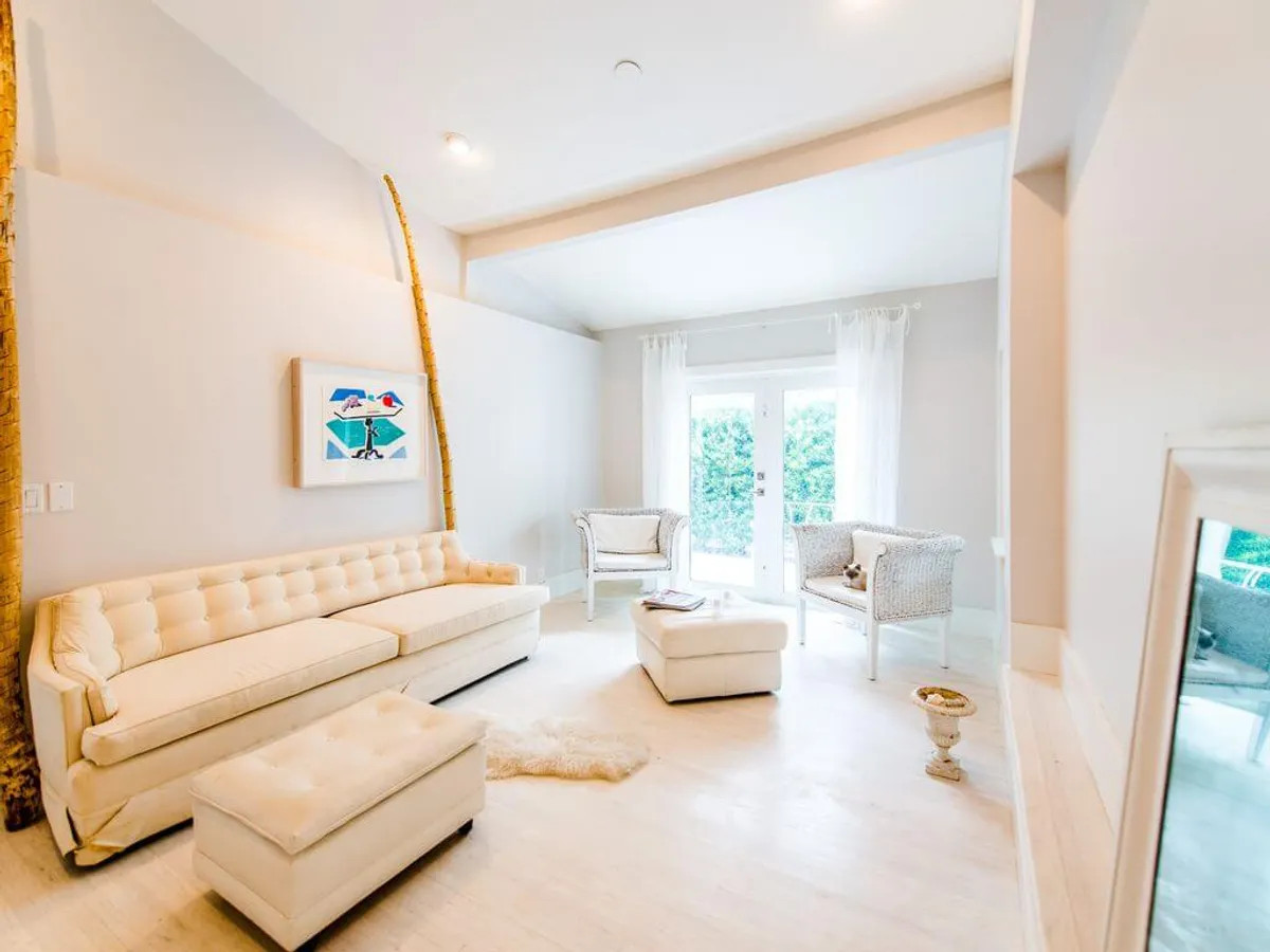 A peaceful, inviting interior scene with a cream tufted sofa, two white wicker chairs, and large French doors adorned with sheer curtains. The doors open to a garden or pool area, and the space is bathed in natural light, creating a calm and social ambiance ideal for gatherings or quiet moments.
