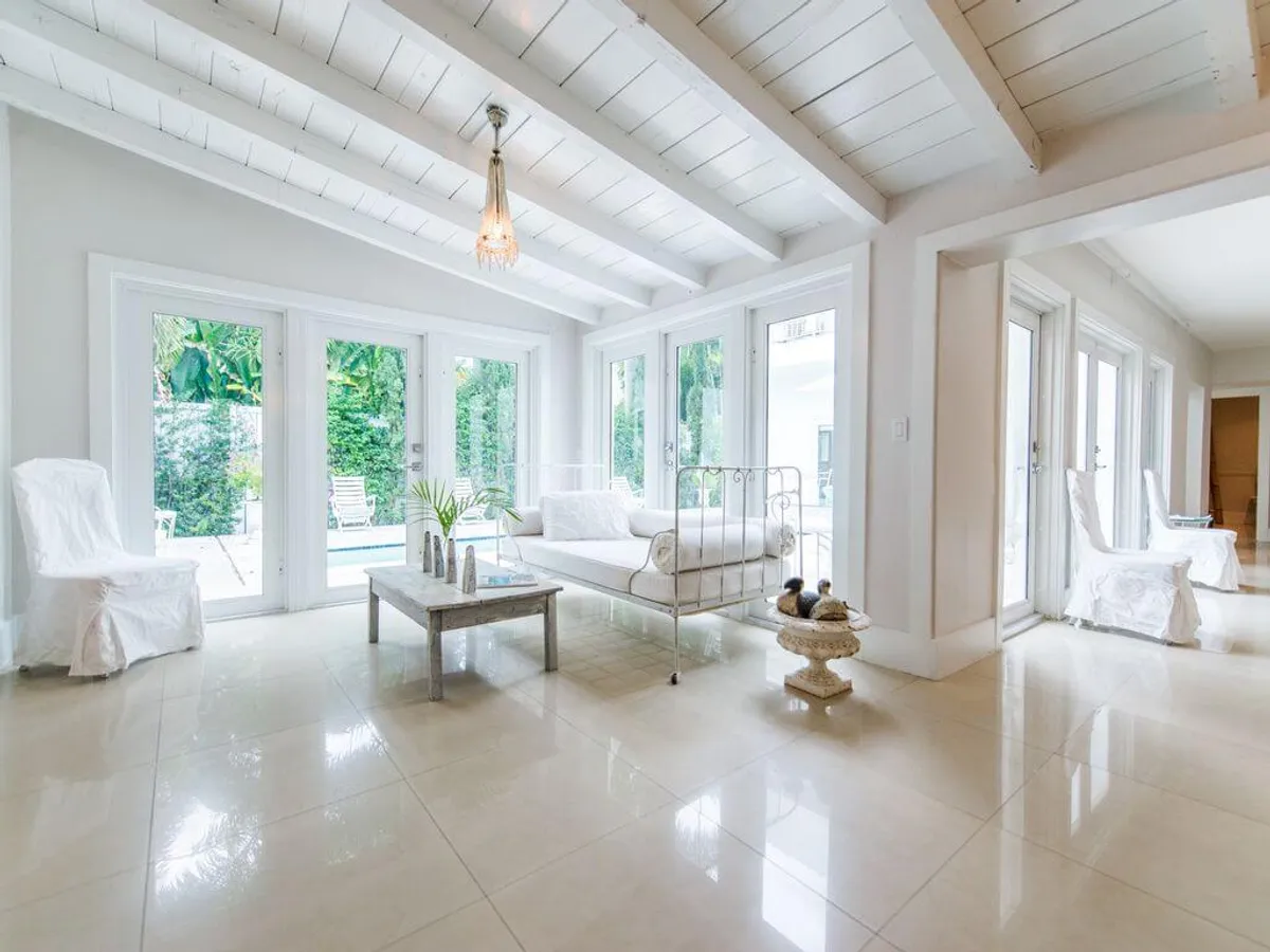 A sunlit living area featuring large French doors and windows that open directly to the pool. The space includes a white wrought iron daybed with soft cushions, a glass-top coffee table with decorative vases, and white slipcovered chairs. Lush greenery and the glimmer of the pool are visible just outside, while natural light fills the room, highlighting the Miami-inspired décor.