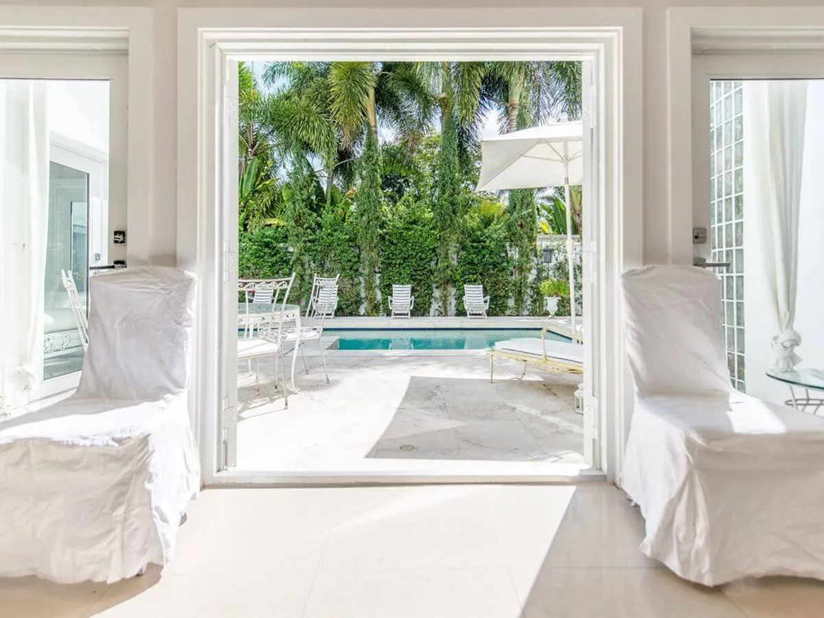 Open French doors from the interior frame a view of the blue pool just outside, with two white slipcovered chairs in the foreground. The lush tropical greenery and palm trees envelop the pool area, blurring the line between indoor comfort and outdoor luxury.