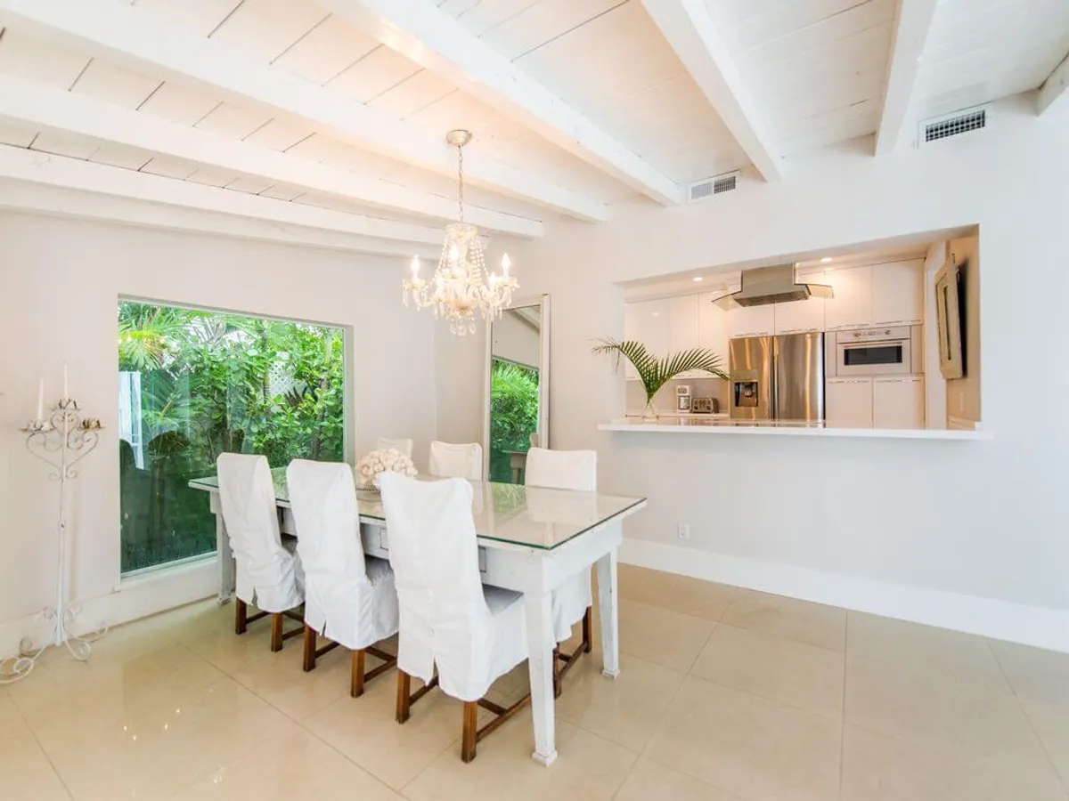 This image showcases a bright dining area with a glass dining table surrounded by white covered chairs. Large windows offer lush tropical garden views, and an elegant chandelier creates a welcoming atmosphere. A pass-through window connects to the modern kitchen, where stainless steel appliances are visible, suggesting a seamless flow for meal preparation and serving.
