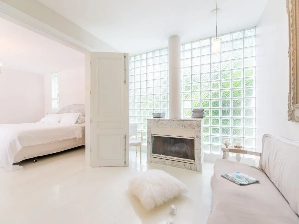 A serene living space highlighted by a large glass block window wall, a marble fireplace, a neutral-toned sofa, and a plush floor cushion. The room is bathed in soft, natural light, offering a peaceful setting for reading, meditation, or quiet reflection, with a sense of privacy and calm.