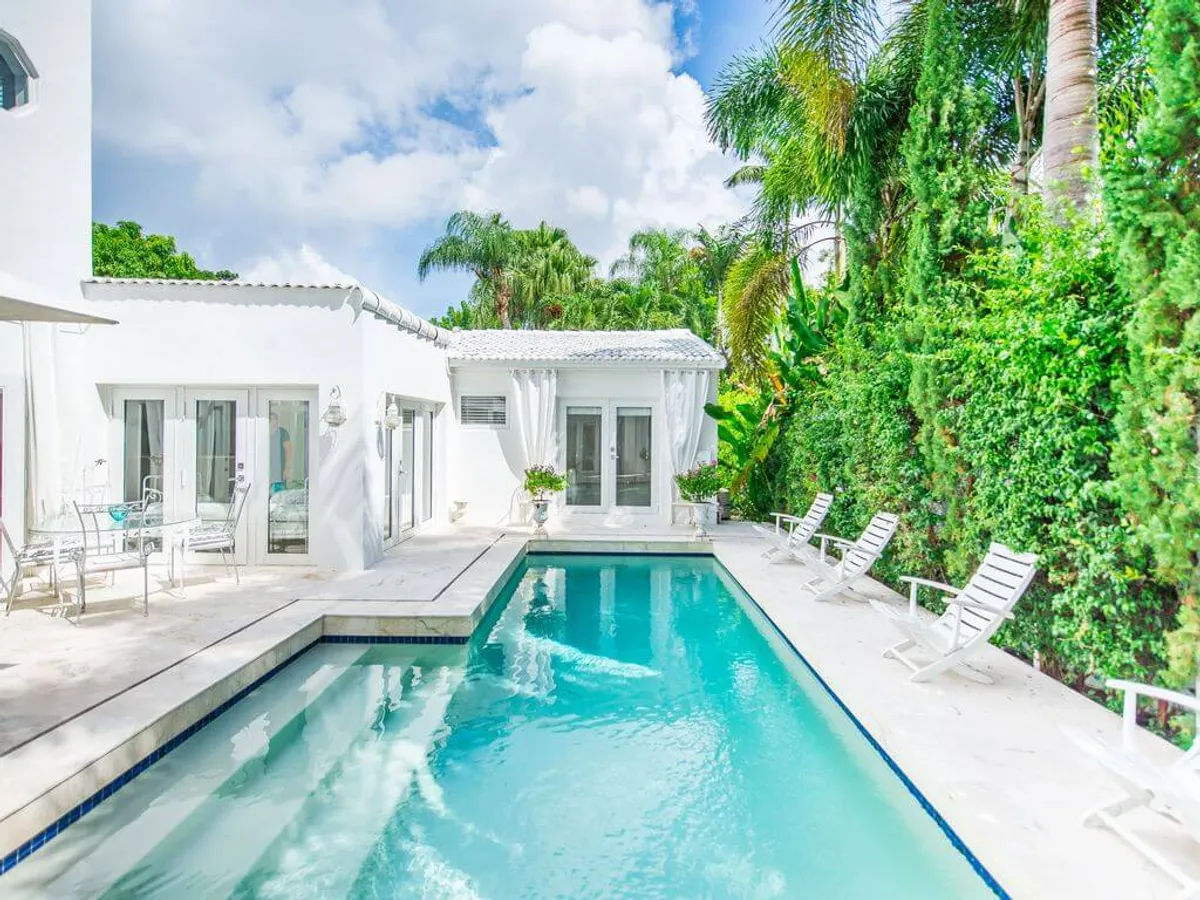 This exterior shot showcases a sparkling heated swimming pool bordered by dense green hedges and tall palms, with a modern white house and multiple French doors opening to the outdoors. Sun loungers and outdoor seating create a relaxing ambiance, reinforcing the connection between indoor comfort and tropical outdoor living.