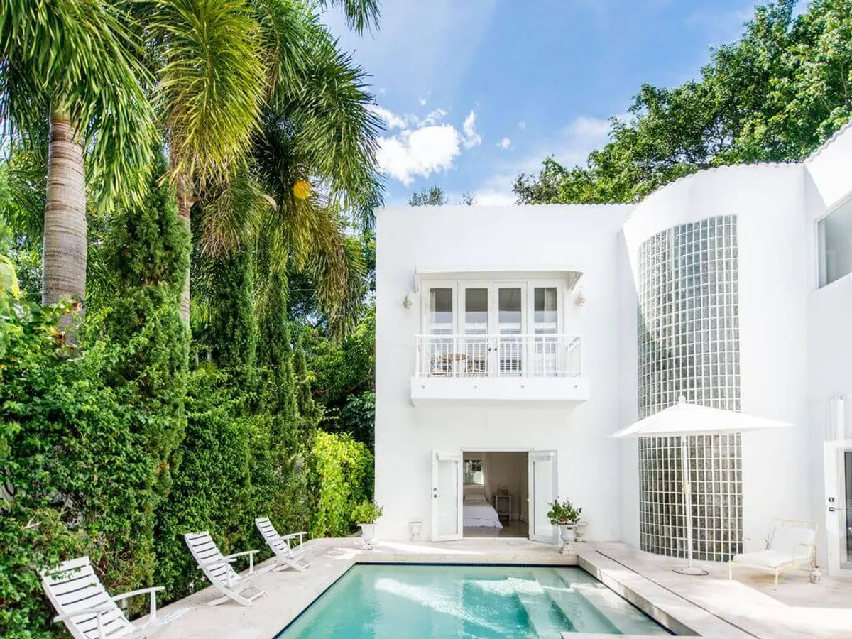 A heated swimming pool with crystal-clear water sits before a modern white house featuring a unique curved glass brick wall and French doors. Tall, lush tropical palms and greenery surround the pool deck, while white balcony railings and poolside chairs enhance the sense of exclusive luxury.