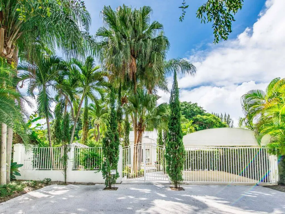 The image presents a welcoming gated entrance flanked by tall cypress trees, with lush palm trees and tropical plants surrounding the curved driveway. The bright blue sky and vibrant greenery evoke a sense of arrival into a private, tropical sanctuary that seamlessly blends with Miami's natural landscape.