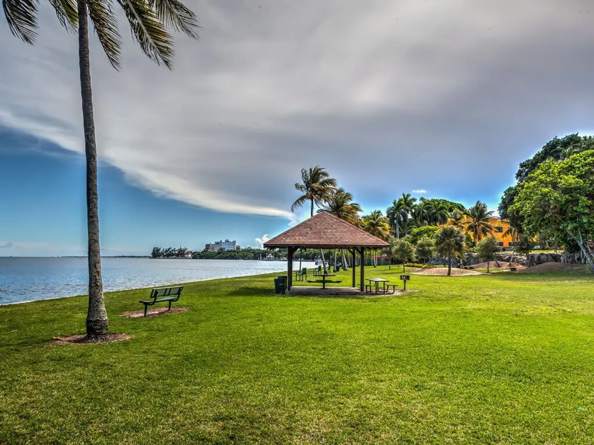 A peaceful waterfront view featuring a pavilion with picnic tables, palm trees, benches facing the water, and people enjoying quiet outdoor activities like picnicking and walking. The scene is serene, inviting visitors to relax by the water while enjoying the tropical breeze.