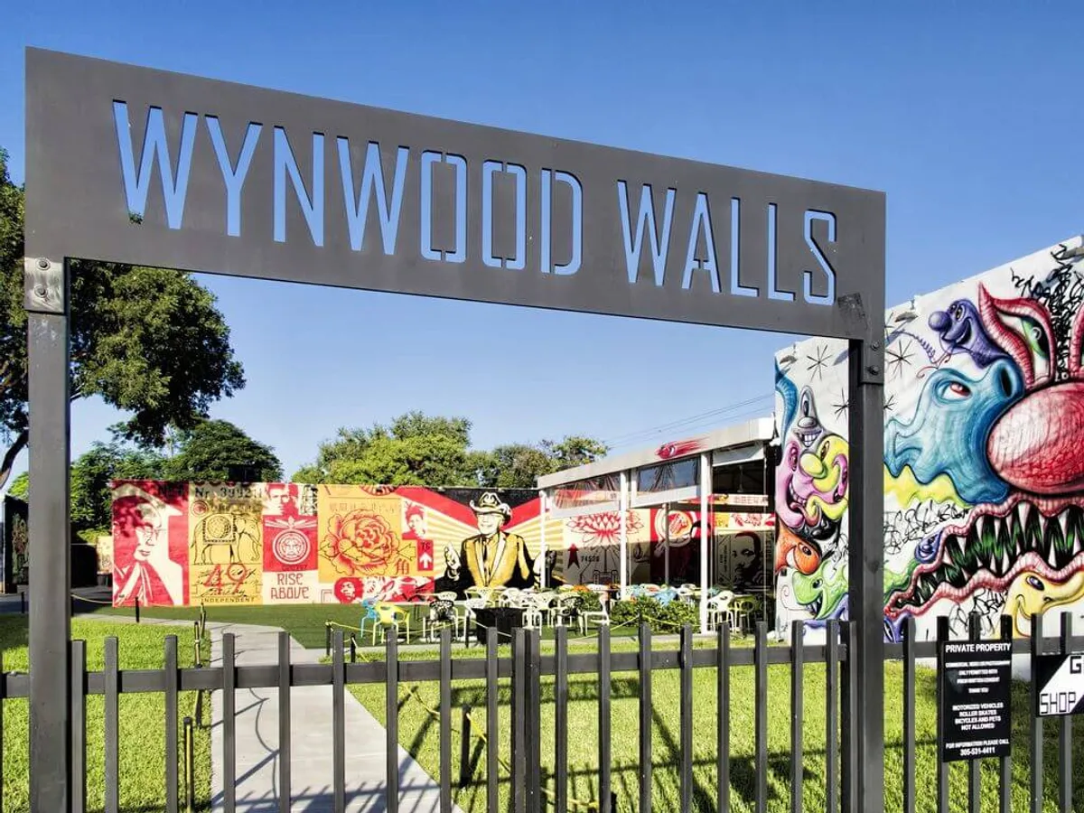 The image captures the vibrant entrance to Wynwood Walls, featuring colorful street art murals that line the walls, an inviting outdoor seating area with tables and chairs, and a well-kept green lawn and walkway welcoming visitors. The atmosphere is lively and artistic, with people gathering to appreciate Miami's dynamic outdoor art scene.