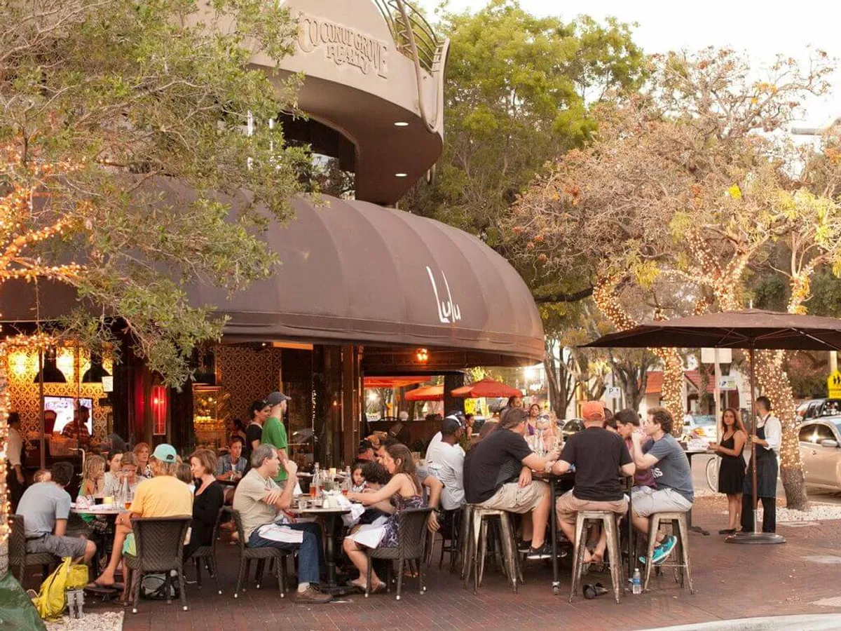 This image captures a lively outdoor dining area with groups of people enjoying meals, a restaurant facade with a distinct curved awning, trees adorned with string lights, and a vibrant street scene featuring pedestrians and parked cars. The overall ambiance is energetic yet relaxed, highlighting the convivial social and dining atmosphere typical of Coconut Grove Village.